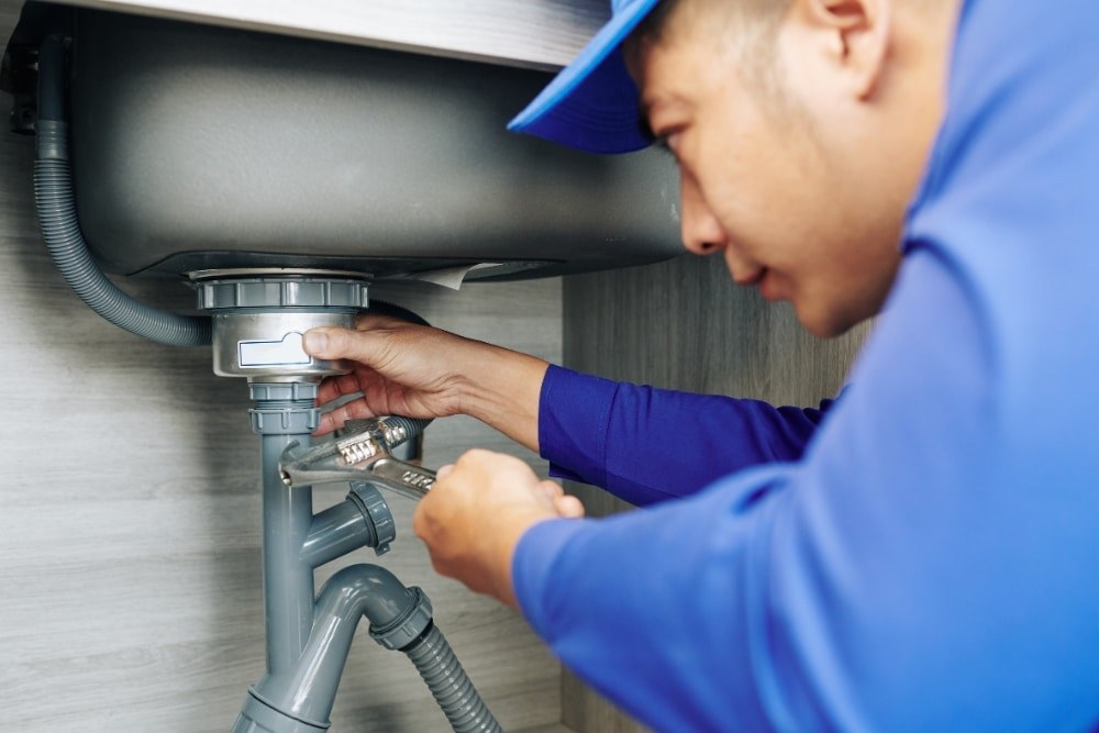 A plumber in a blue uniform dismantling the trap under a stainless steel kitchen sink