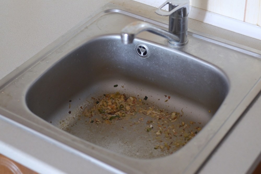 Food scraps and sediment left sitting around the waste outlet of a stainless steel kitchen sink