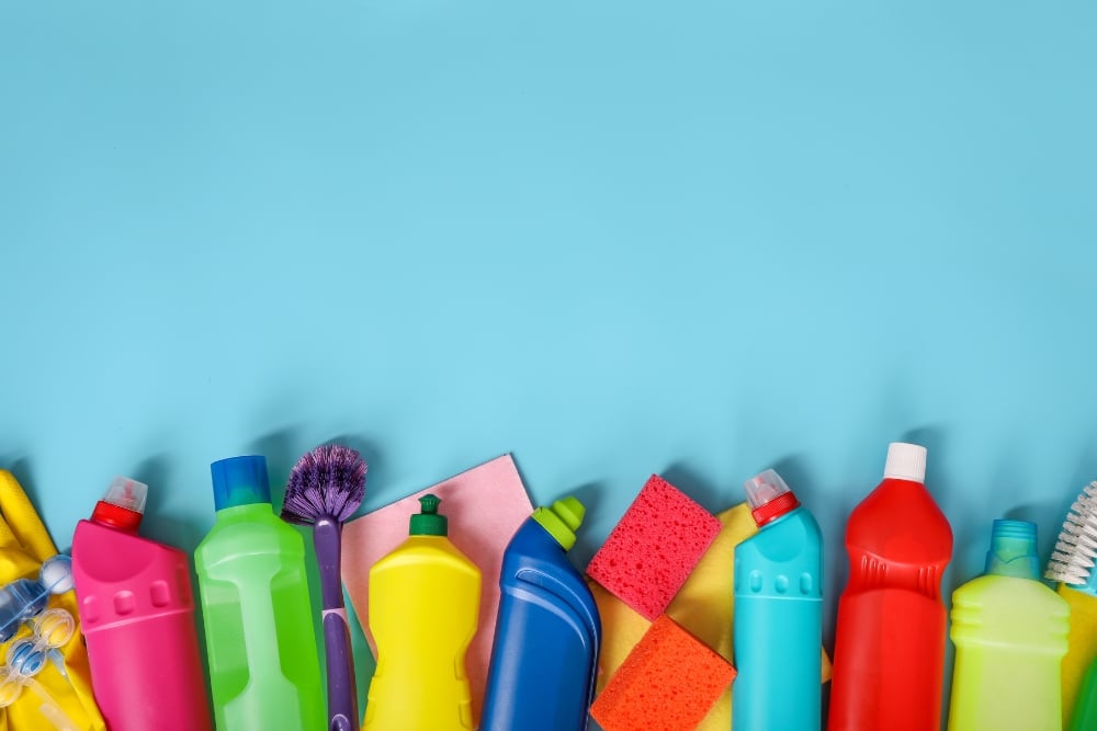 A row of household cleaning product bottles and sponges lined up against a blue background